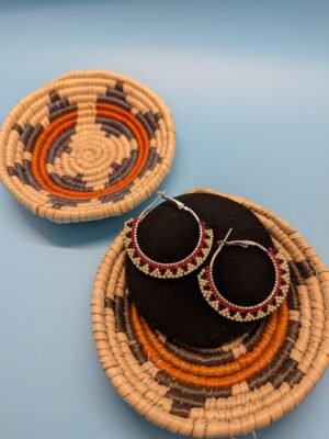 Mesa Steps hoop earrings by Adakair, featuring a striking geometric pattern of cream, black, and red beads. The earrings are shown on a display stand with a woven basket in the background.